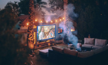 Screen on side of house surrounded by fairy lights showing video and facing set of couches around a fire