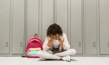 A young girl sits in a school hallway, holding her head in her hands. 