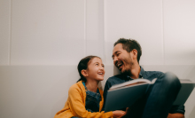 A father and daughter sit against a white wall reading a book. They are looking at each other and laughing.