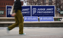 An out of focus college student walks by a series of blue and white signs that read "Judge Janet Protasiewicz Wisconsin Supreme Court".
