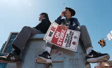 Two young people sit on top of a steel box. The one on the right dangles a red and white signs that reads, "We R on strike for our students and communities". 