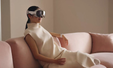 An Asian woman sits on a light pink couch, She is in a white dress and is wearing the Vision pro on her head.