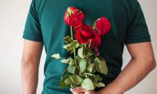 A man wearing a teal T-shirt holds a bunch of wilted red roses behind his back.