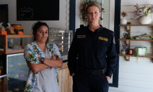 Two women, a tall one in a dark blue Australian police uniform and a short one in a tropical shirt, standing in a bakery.
