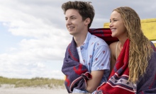 A young man and a woman sit together on a beach, sharing a beach towel.