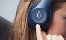 Woman in side profile with Beats Studio3 headphones on and a finger up to one of her ears.