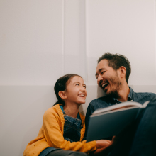 A father and daughter sit against a white wall reading a book. They are looking at each other and laughing.