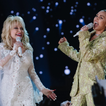 Dolly Parton wears a glittering white outfit and sings next to Miley Cyrus, who is wearing a green lace top and belting into a microphone.