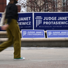 An out of focus college student walks by a series of blue and white signs that read "Judge Janet Protasiewicz Wisconsin Supreme Court".