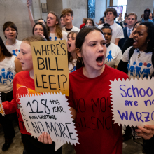 A group of young people in red and white shirts march and chant through a legislative building. They hold signs reading, "Schools are not warzones", "Ban assault weapons", "Where's Bill Lee?", and "128  mass shooting this year. No more."