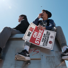 Two young people sit on top of a steel box. The one on the right dangles a red and white signs that reads, "We R on strike for our students and communities". 