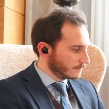 man is sitting down, looking to his left, with translator earbuds in his right ear 