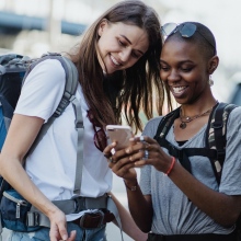 Two smiling women with backpacks look at a mobile phone.