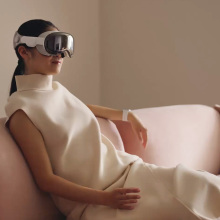 An Asian woman sits on a light pink couch, She is in a white dress and is wearing the Vision pro on her head.