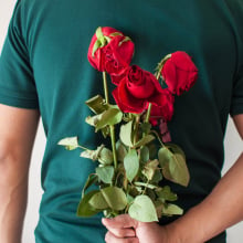 A man wearing a teal T-shirt holds a bunch of wilted red roses behind his back.