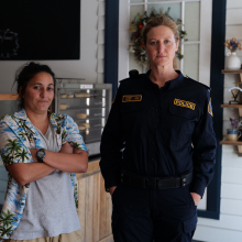 Two women, a tall one in a dark blue Australian police uniform and a short one in a tropical shirt, standing in a bakery.