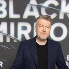A man with silverish hair and a light beard stands in front of a step-and-repeat for "Black Mirror."