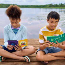 Two kids reading from a Kindle Paperwhite Kids e-reader while sitting on a dock