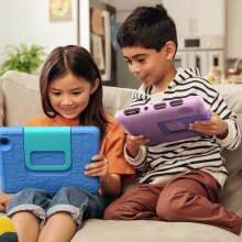 Two kids sitting on the couch and playing on their Amazon Fire 7 Kids tablets with smiles on their faces.