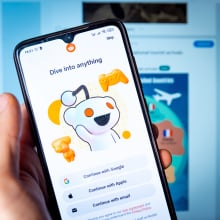 Reddit welcome page depicted on a smartphone being held by a hand in front of a computer monitor