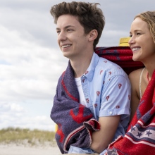 A young man and a woman sit together on a beach, sharing a beach towel.