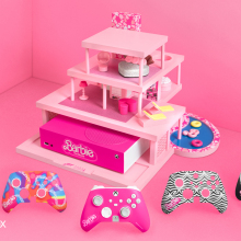 A product photo of the 5 new Barbie XBox controller skins in the foreground. In the background, a Barbie-themed xbox (bright pink, with the Barbie logo) housed in a mini version of Barbie's dream house, complete with pool.