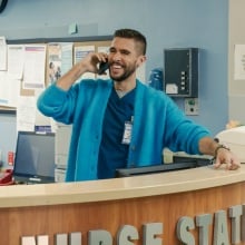 Lance from "The Other Two" takes a call at a hospital desk while wearing blue scrubs and a blue cardigan.