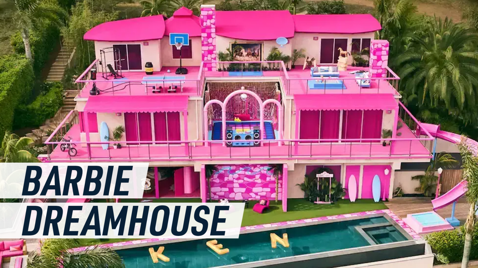 Drone aerial shot of the pink Barbie Dreamhouse, gold balloon letters spelling KEN float in the swimming pool.