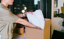 a woman in a tan polo packs a stack of white t-shirts into a cardboard box. there are four beaded bracelets on her wrist