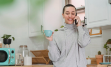 a woman with a shaved head wearing a light gray sweater talks on a smartphone in her kitchen while holding a blue teacup