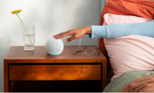 a person in a blue shirt touches the top of a fifth-gen echo dot (with clock) that's sitting on a wooden bedside table next to a pair of glasses and a vase with a yellow flower