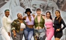The six influencers stand with their arms around each other. In the background is an enlarged photo of stacked Shein packages.