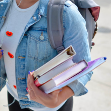 a woman in a denim jacket carries a stack of books and binders while wearing a gray backpack