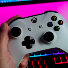 a close-up of a person holding an xbox controller in front of.an RGB keyboard and a gaming monitor