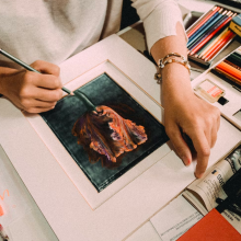 a close-up of a woman drawing a picture of a rose with colored pencils on a messy art desk