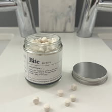 Bite Toothpaste jar with lid off and Bits on countertop with faucets and toothbrushes in background
