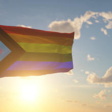 The Progress Pride flag, featuring rainbow stripes and a triangle of pink, blue, white, brown, and black stripes, waves from a flag pole over a sunny sky background