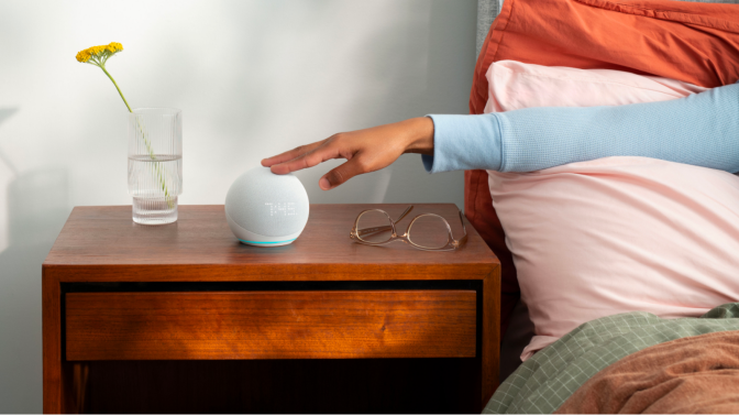 a person in a blue shirt touches the top of a fifth-gen echo dot (with clock) that's sitting on a wooden bedside table next to a pair of glasses and a vase with a yellow flower